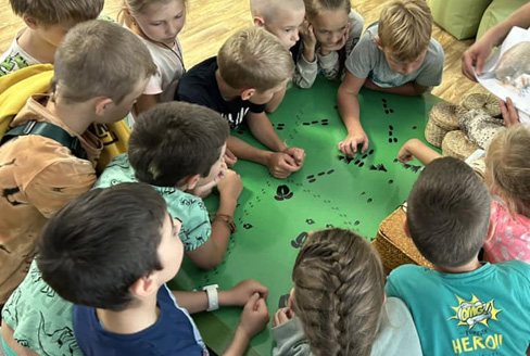 Bērni ap galdu, uz kura zaļš plakāts ar melniem, zīmētiem dzīvnieku pēdu nospiedumiem.